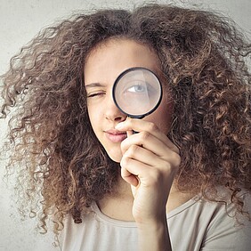 Eine Person mit lockigem Haar hält eine Lupe vor ihr Auge. Die prüft und möchte es ganz genau wissen.nd es sind keine weiteren sichtbaren Elemente oder Texte im Bild.