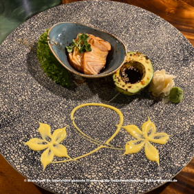 Das Bild zeigt ein kunstvoll angerichtetes Gericht mit Lachs in einer blauen Schale, einer halben Avocado mit Gewürzen, einer weißen blumenförmigen Garnitur und grüner Paste (vermutlich Wasabi). Der Teller ist mit gelben, blumenartigen Designs dekoriert.